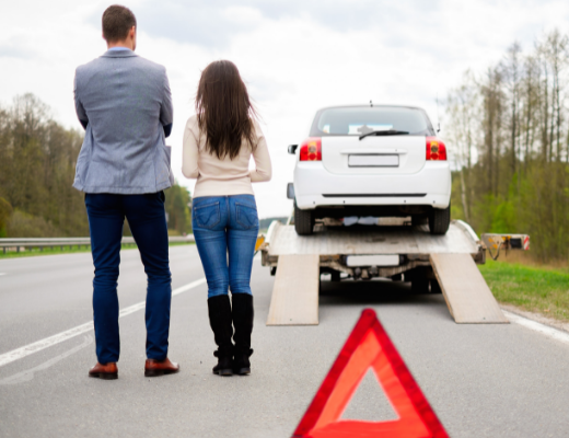 Man and woman standing on the side of the road watching their car be loaded up onto a truck.